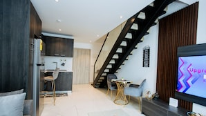 A modern living room with a flat-screen TV, a dining area with a round table and chairs, a staircase, and a kitchen with dark cabinetry. - The Lennox 802 One Bed Duplex (Accra)