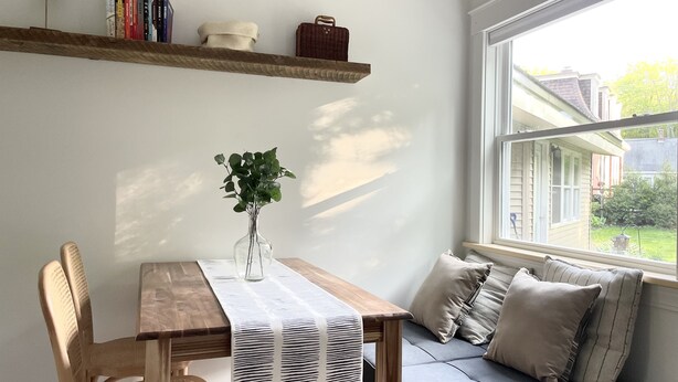Une pièce chaleureuse avec une table en bois, un banc et une fenêtre donnant sur une maison et de la verdure.