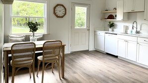 Une cuisine avec des meubles blancs, une table à manger en bois avec deux chaises, une horloge au mur et une fenêtre donnant sur un paysage verdoyant.