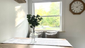 Un coin repas avec une table en bois, un vase en verre transparent contenant des plantes vertes et une vue sur les arbres par la fenêtre.