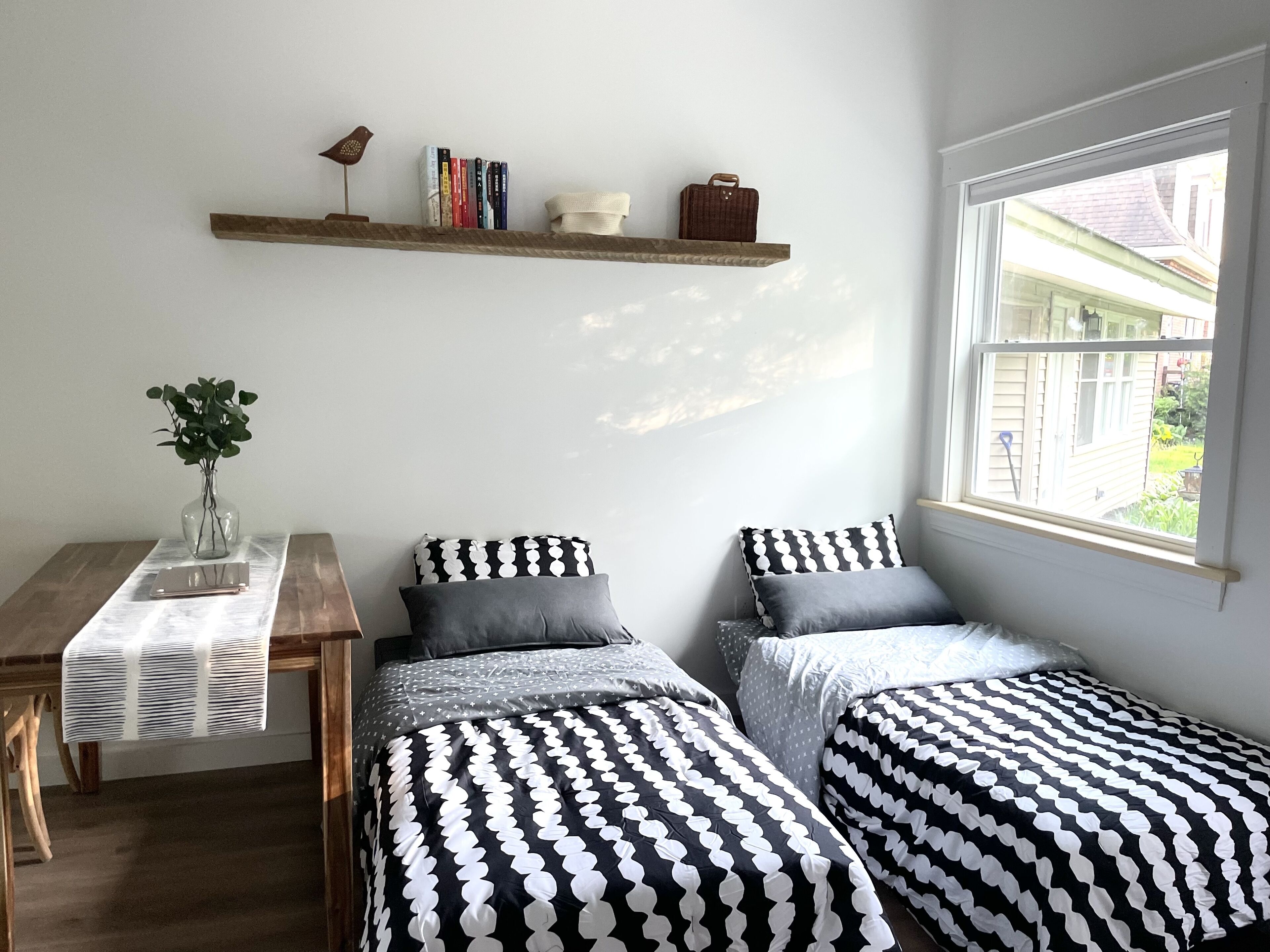 Una camera da letto con due letti, un comodino con una pianta, una finestra vista e uno scaffale con libri e una lampada.