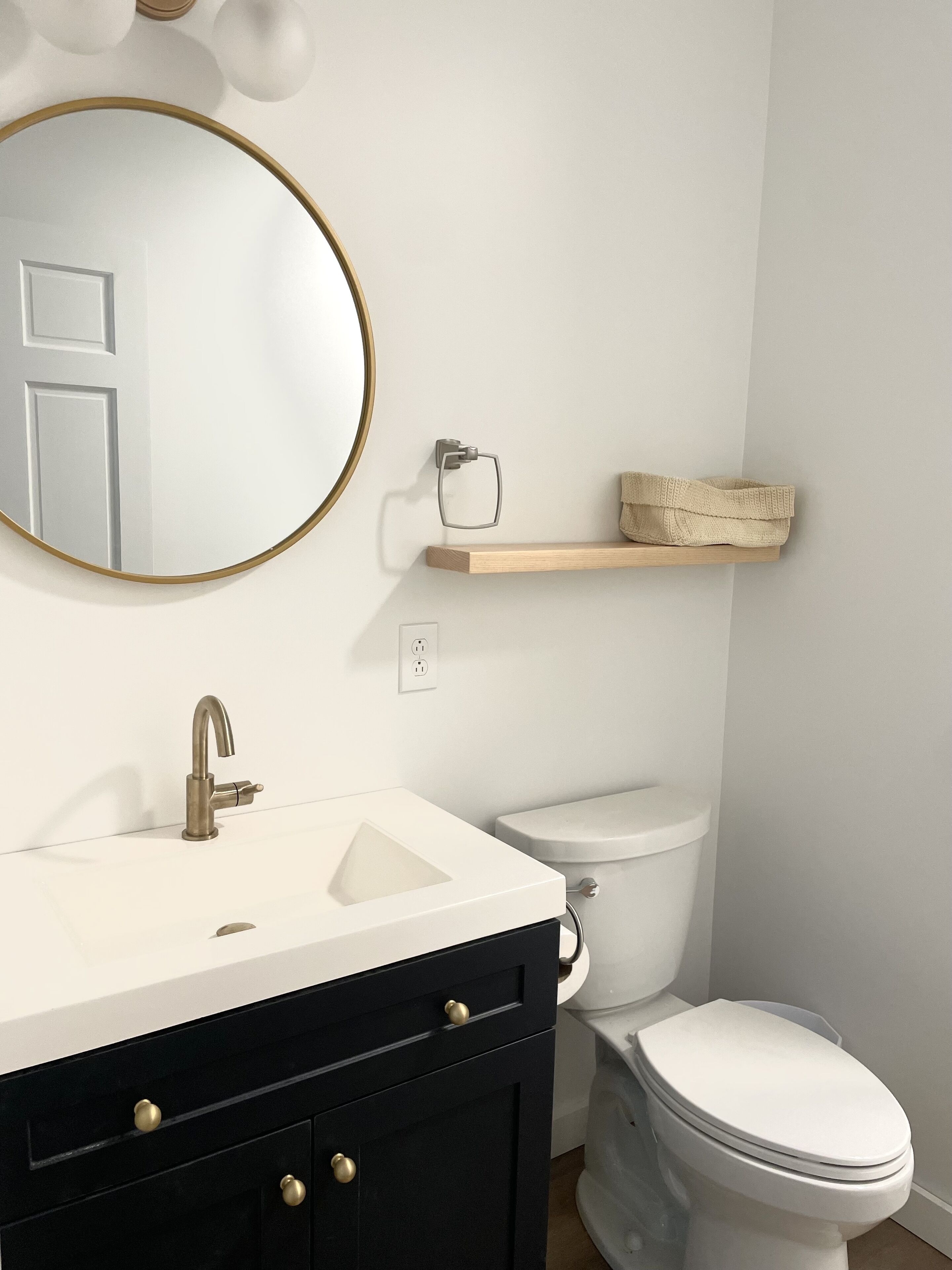 A bathroom with a white sink, a round mirror, a toilet, and a wooden shelf.