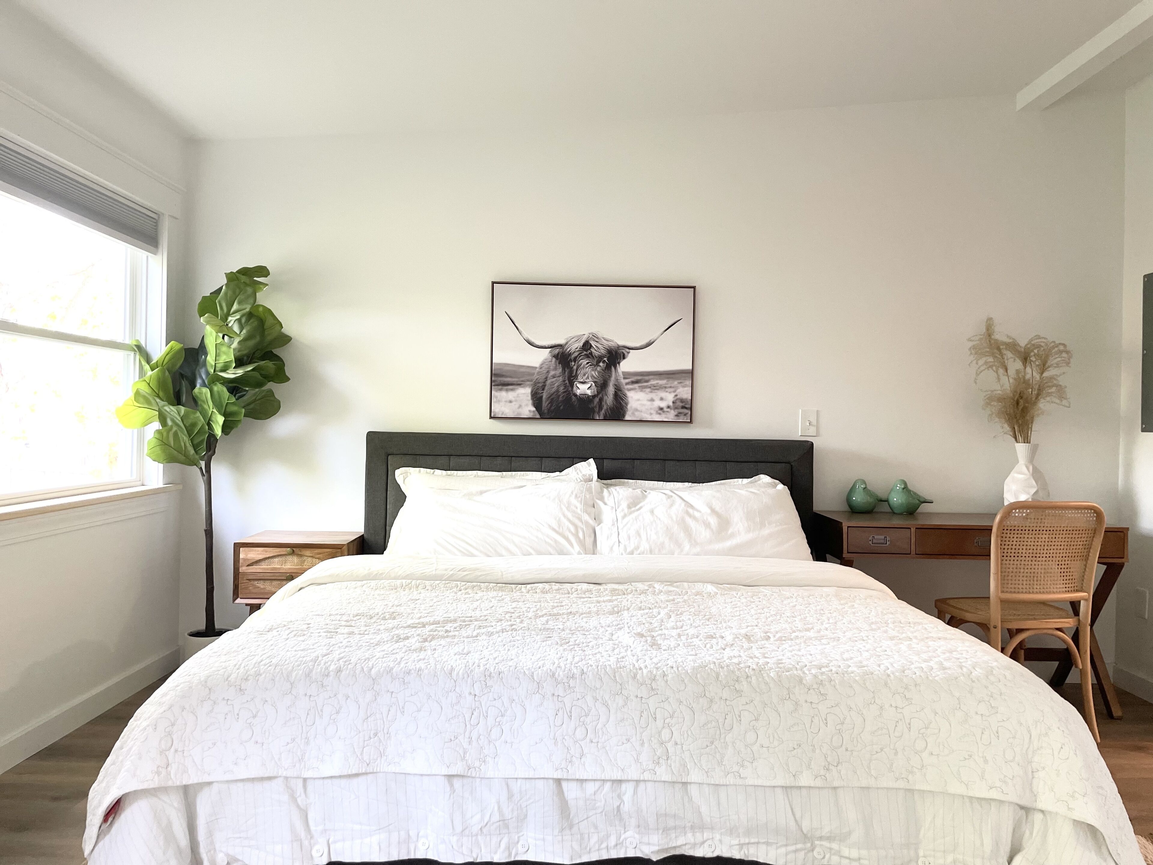 Une chambre à coucher avec un grand lit, une tête de lit, une table de chevet, une commode, une fenêtre, une plante, un tableau accroché au mur et une chaise.