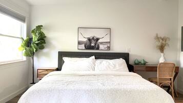 Une chambre à coucher avec un grand lit, une tête de lit, une table de chevet, une commode, une fenêtre, une plante, un tableau accroché au mur et une chaise.
