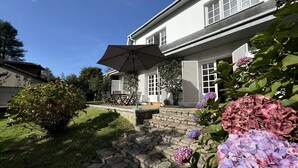 Een huis met een stenen pad dat leidt naar een patio met een tafel en stoelen, omgeven door weelderige begroeiing en hortensia's.