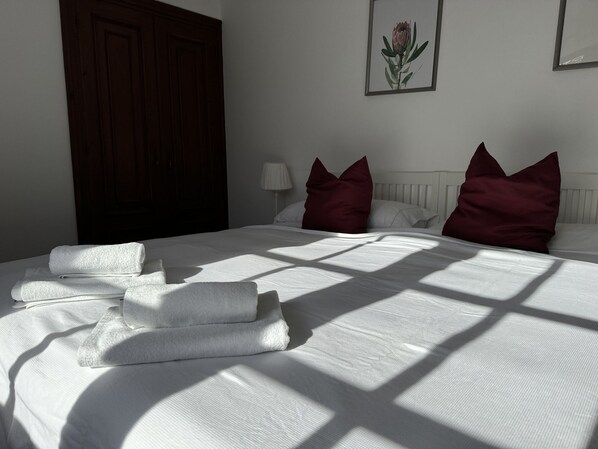 Un letto rifatto con lenzuola bianche e asciugamani piegati, un armadio in legno e una lampada a parete.