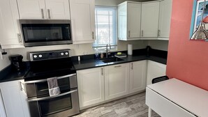 A modern kitchen with white cabinets, a black stove, and a microwave. There's a window with blinds, a sink, and a small table.