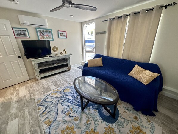 A living room with a blue sofa, a glass coffee table, a TV on a wooden stand, and a patterned rug.