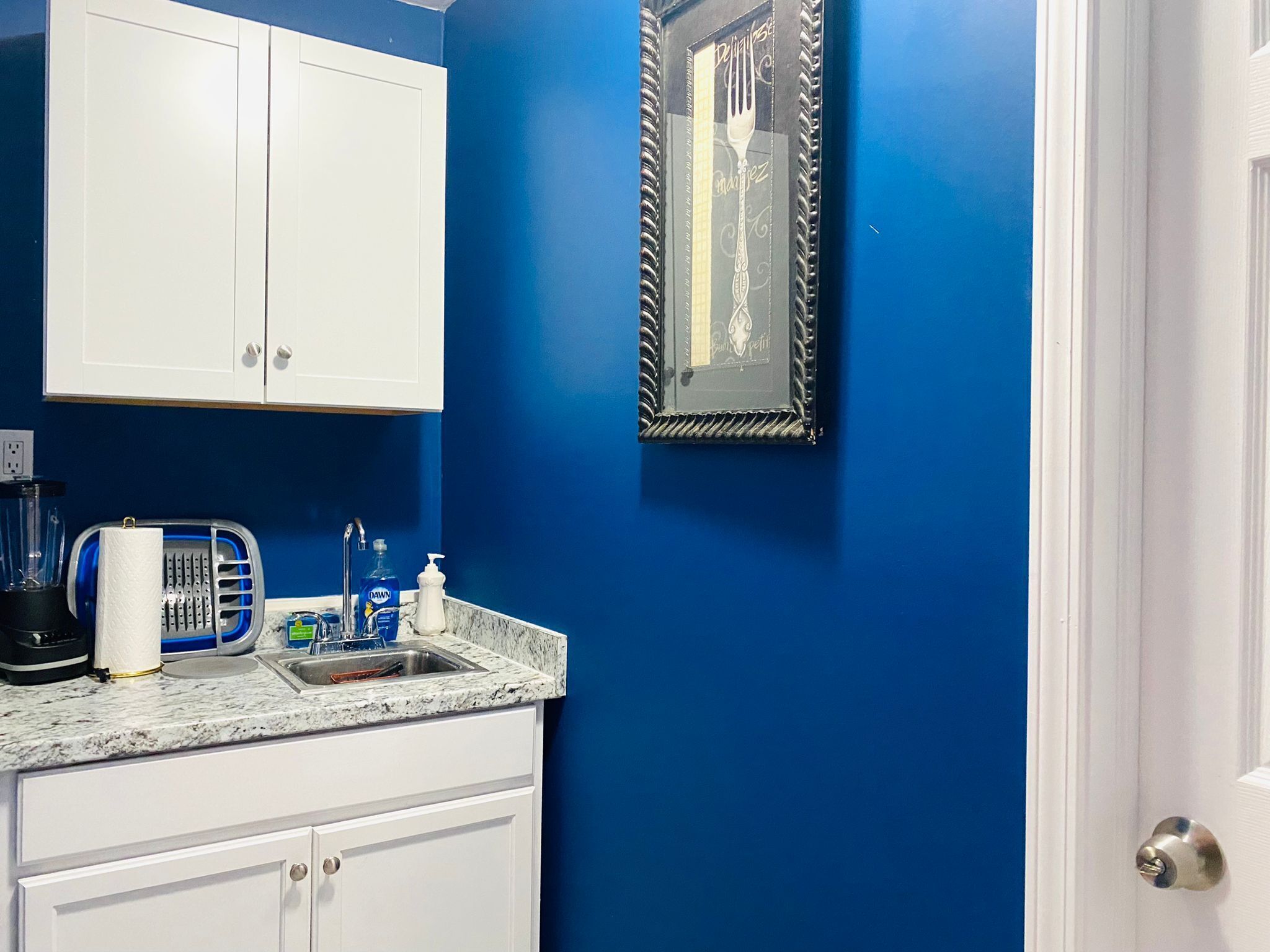 Una cocina con armarios blancos, pared azul, un lavamanos y un cuadro en la pared.