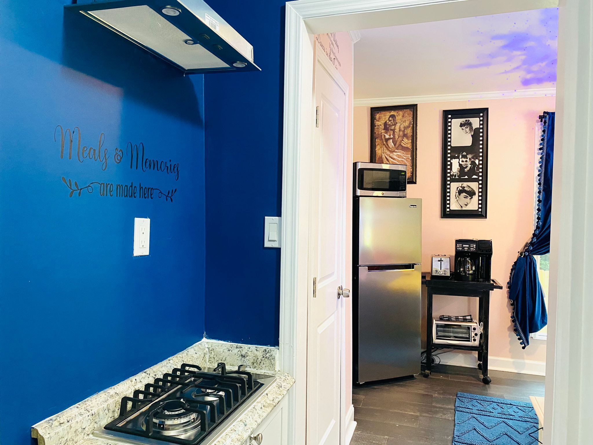 Une cuisine avec des murs bleus, une cuisinière à gaz, un réfrigérateur et un four à micro-ondes. Une porte donne accès à une autre pièce où se trouvent une machine à café et un tableau encadré accroché au mur.