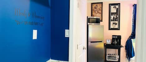 A kitchen with blue walls, a gas stove, a refrigerator, and a microwave. There is a door leading to another room with a coffee maker and a framed picture on the wall.
