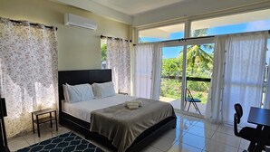 A bedroom with a bed, a desk, and a view of greenery outside. - Stylish modern apt beach view (Belmont)