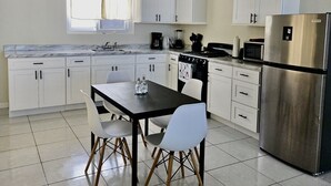 A modern kitchen with white cabinets, a stainless steel refrigerator, a dining table with chairs, and a window with curtains. - Stylish modern apt beach view (Belmont)