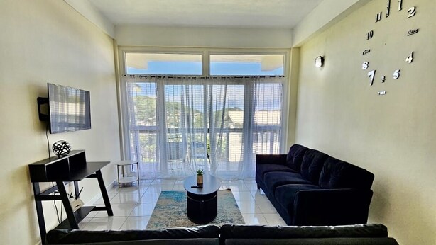 A modern living room with a black sofa, a coffee table, a TV on a stand, and a large window with a view. - Stylish modern apt beach view (Belmont)