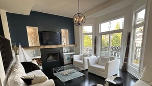 A modern living room with a fireplace, white sofas, a glass coffee table, and large windows offering a view of the outdoors. - Waterfront home w/beach, pool, and tennis. 5 min to Blue Mountain.  (Collingwood)