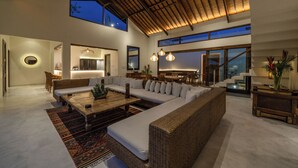 Ein geräumiges Wohnzimmer mit einer großen Eckcouch, einem Couchtisch und einem Essbereich mit Stühlen. Im Hintergrund befindet sich eine Küche, und der Raum verfügt über große Fenster und eine hohe Decke.