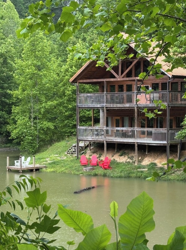 A multi-story wooden cabin with a balcony, situated by a lake surrounded by dense greenery.