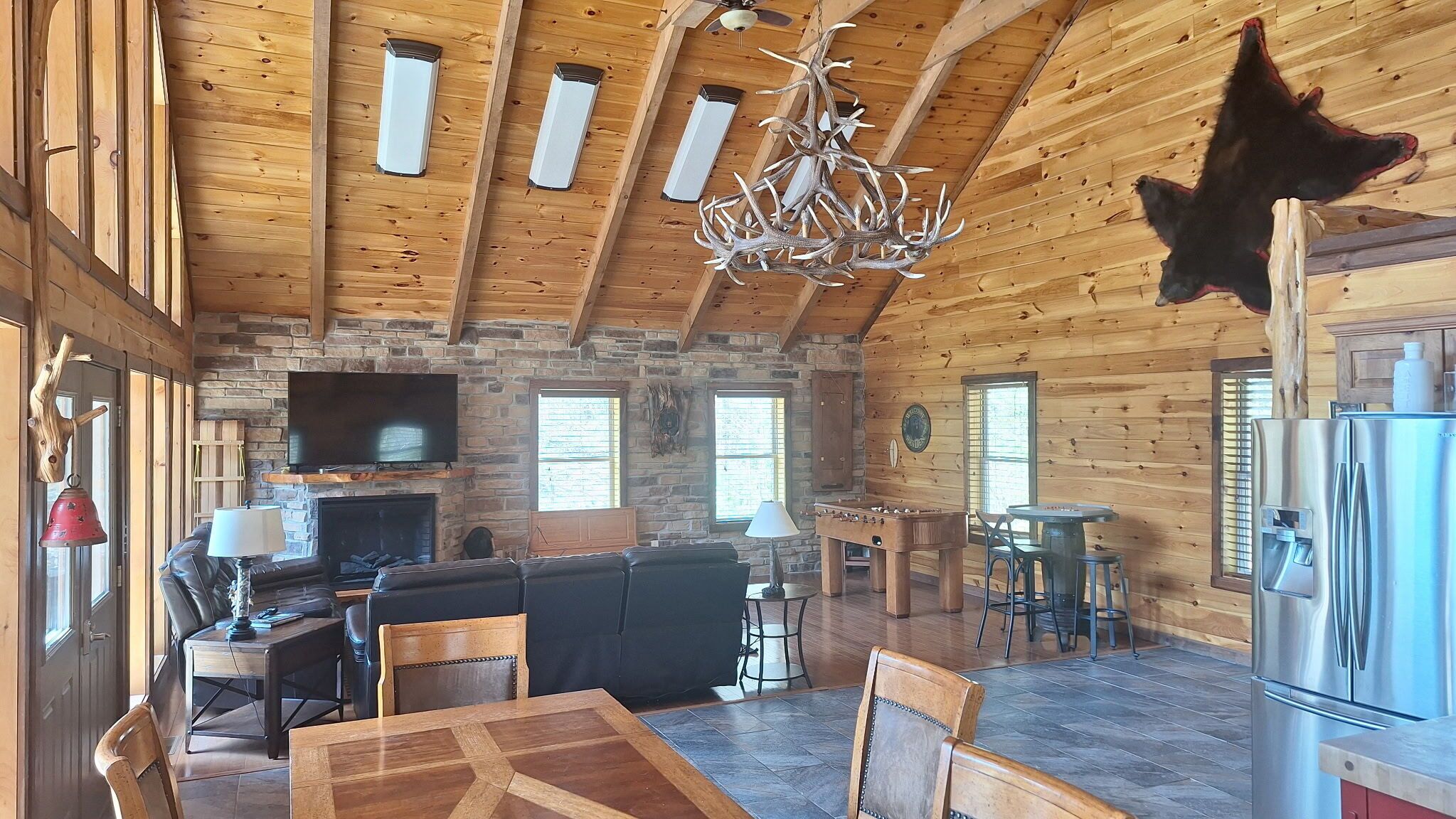 A spacious wooden cabin interior with a stone fireplace, a television, a dining area, and a kitchen with a refrigerator.