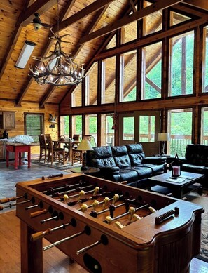 A spacious room with a wooden ceiling, large windows, a leather sofa, a foosball table, and a chandelier.