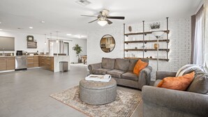 Ein geräumiger Wohnbereich mit grauem Sofa, Hocker und Couchtisch. Im Hintergrund befindet sich ein Deckenventilator, ein Kamin und eine Küche.