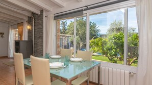 A dining area with a glass table, beige chairs, and a view of a garden through large windows.
