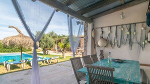 A covered outdoor dining area with a table and chairs, a pool in the background, and fishing nets hanging on the wall.