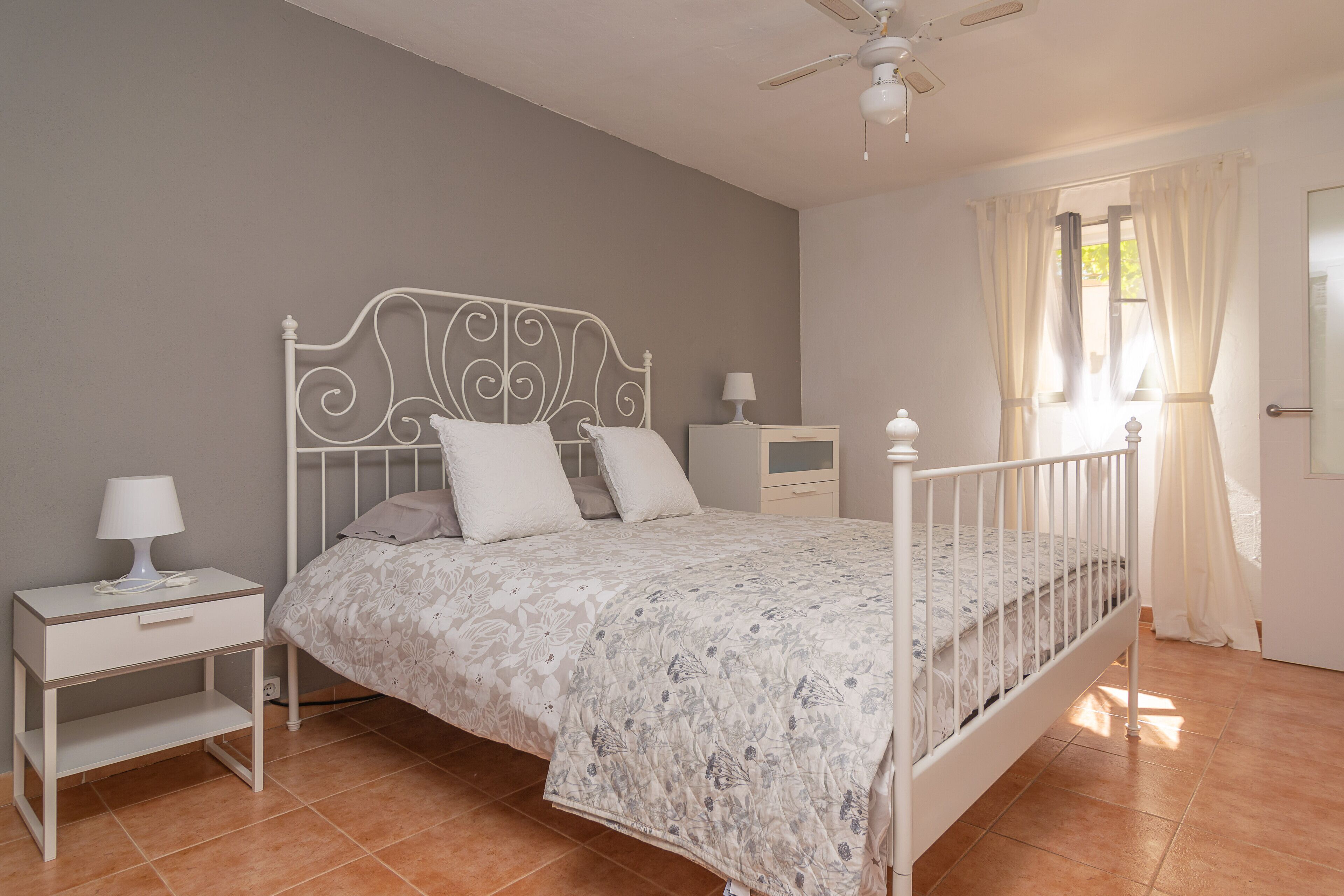 Een slaapkamer met een bed met een wit metalen frame, een bloemenbeddengoed, een nachtkastje met een lamp en een nachttafeltje.
