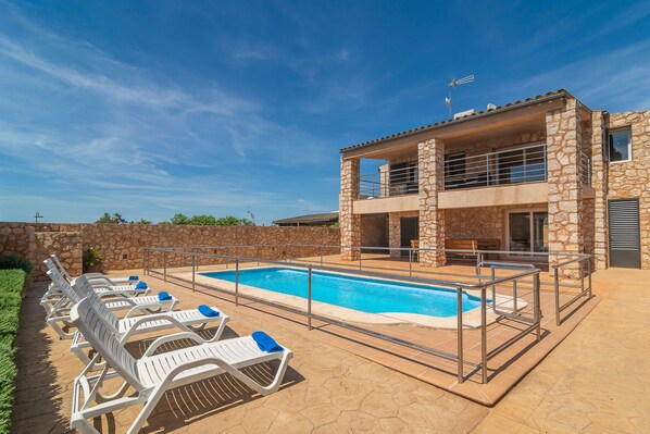 Un espace aménagé au bord de la piscine, avec des chaises longues et un bâtiment en pierre doté d’un balcon.