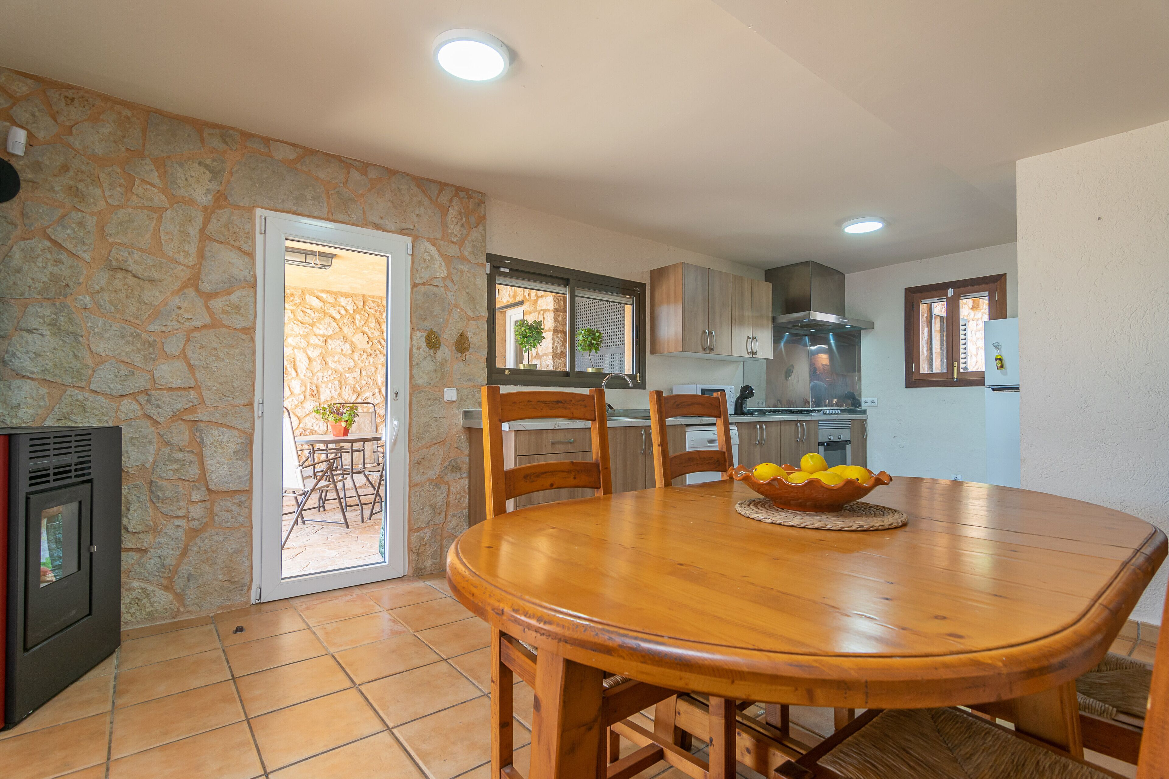 Un comedor con mesa y sillas de madera, una pared de piedra y una cocina al fondo.