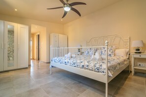 Une chambre à coucher avec un lit en métal blanc, un ventilateur de plafond, une table de chevet avec une lampe et une armoire.