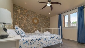 A bedroom with a stone wall, a bed with a floral bedspread, a ceiling fan, a bedside table with a lamp, and a window with curtains.