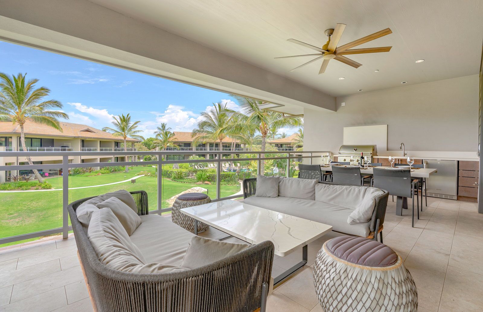 A spacious balcony with a modern outdoor seating area, a kitchen, and a view of a lush garden and pool.