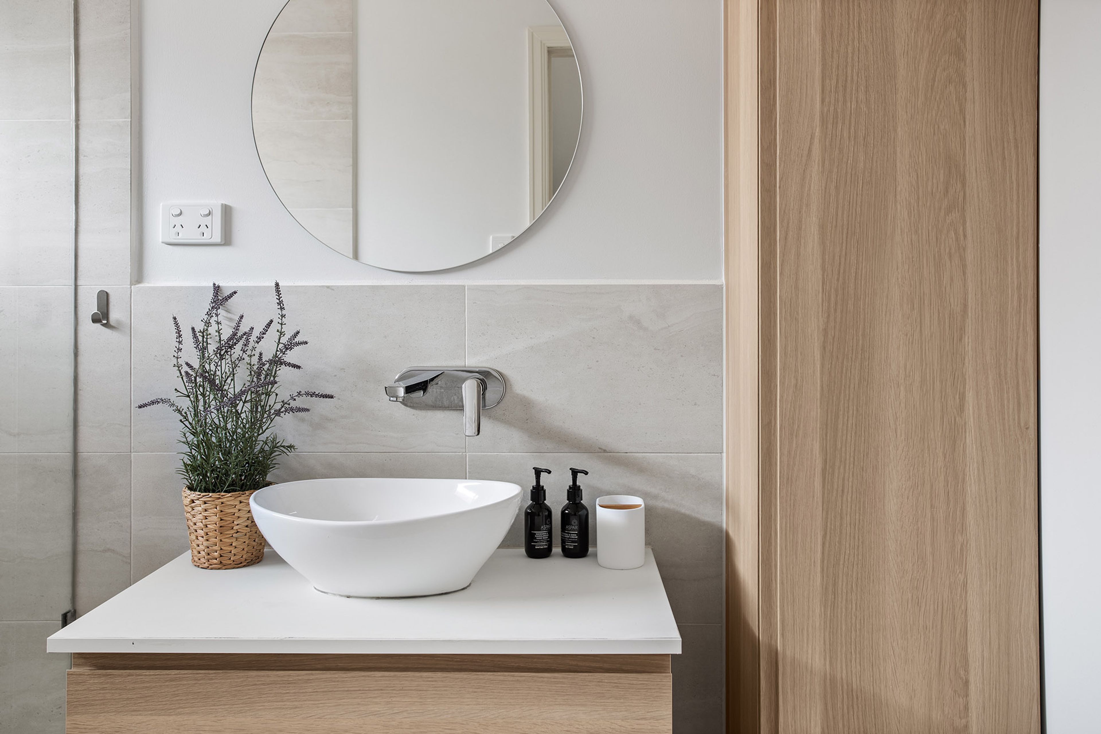 A bathroom with a white sink, a round mirror, a potted plant, and toiletries on the countertop.