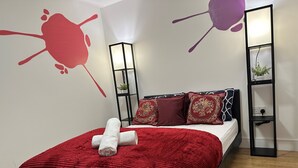 A neatly made bed with a red blanket and two pillows, a small bedside table with a plant, and a wall with a red abstract design.