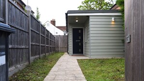 A narrow stone pathway leading to a small shed with a dark door, surrounded by a wooden fence and a grassy area.