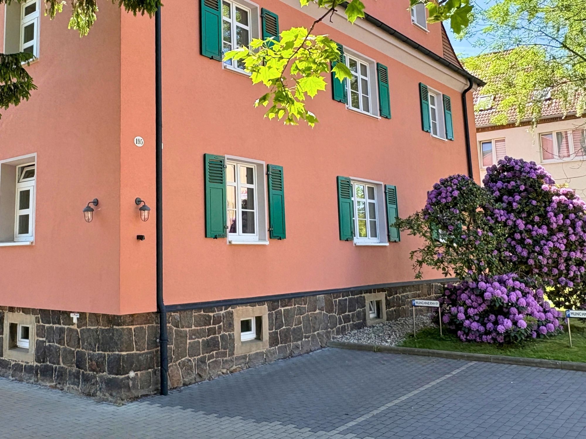 Ein zweistöckiges Gebäude mit grünen Fensterläden, einer Steinsockel und einem gepflasterten Eingang.