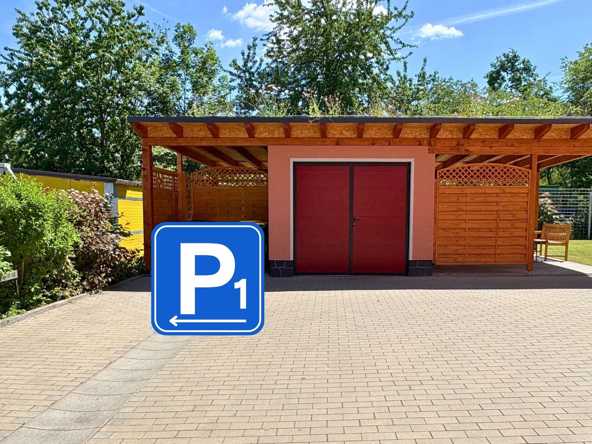 Ein überdachter Parkbereich mit roter Tür, blauem Piktogramm und Pflasterweg.