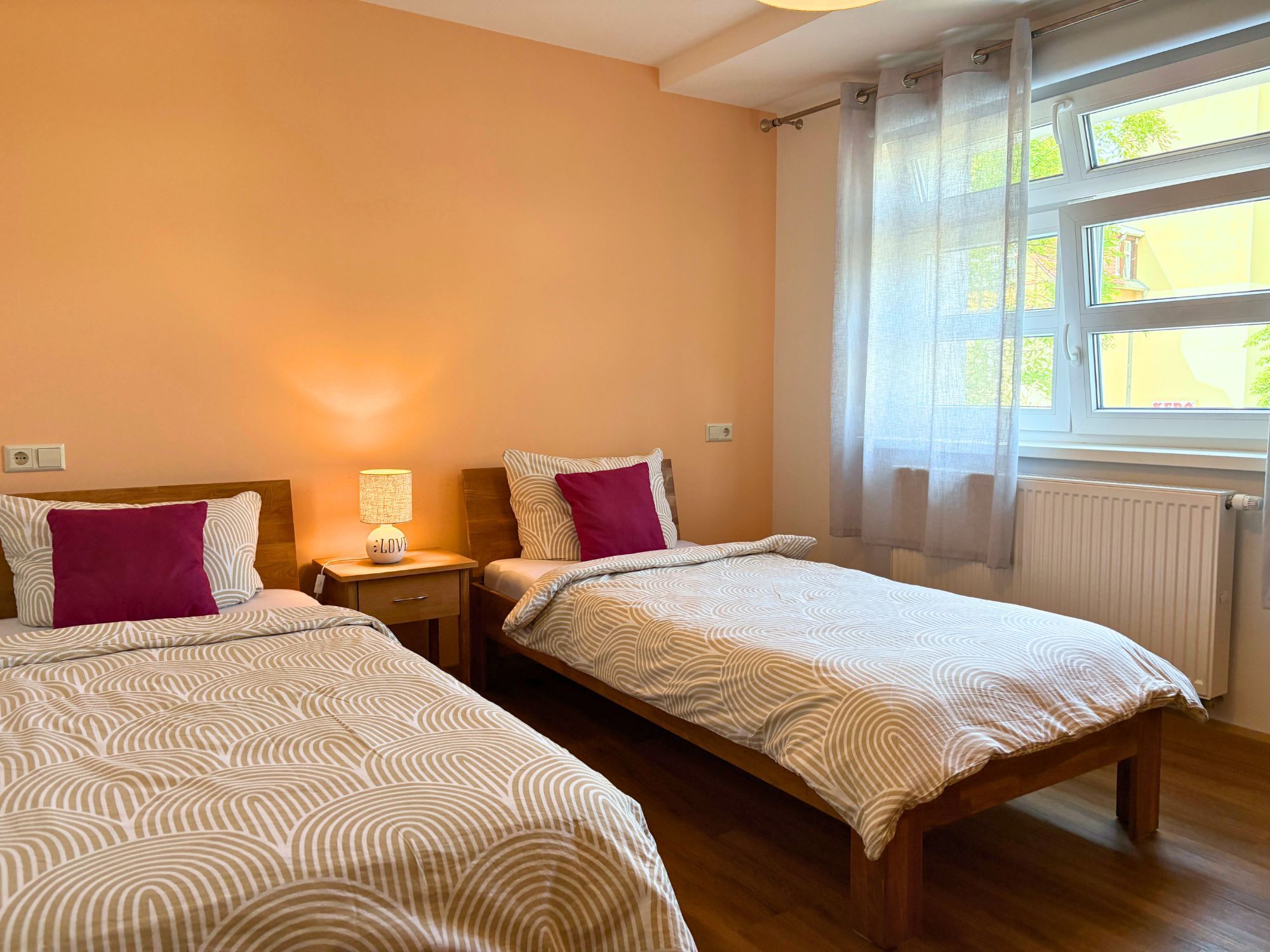 Ein Schlafzimmer mit zwei Einzelbetten, einem Nachttisch mit Lampe, einem Fenster mit Vorhängen und einer Heizung.