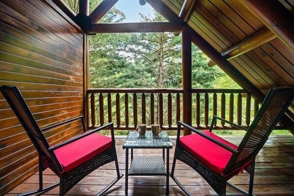 Eine Holzterrasse mit zwei Stühlen und einem Tisch, umgeben von einer Holzbrüstung, mit Blick auf ein bewaldetes Gebiet.
