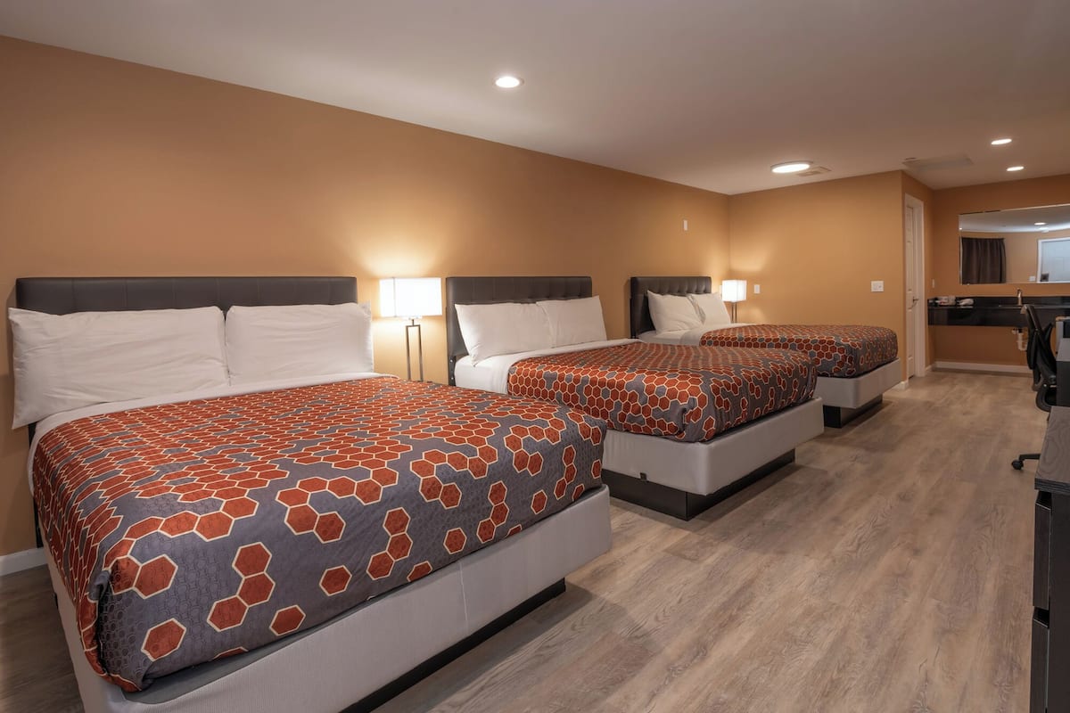 A hotel room with three beds, each with a patterned bedspread, bedside lamps, and a wooden floor.