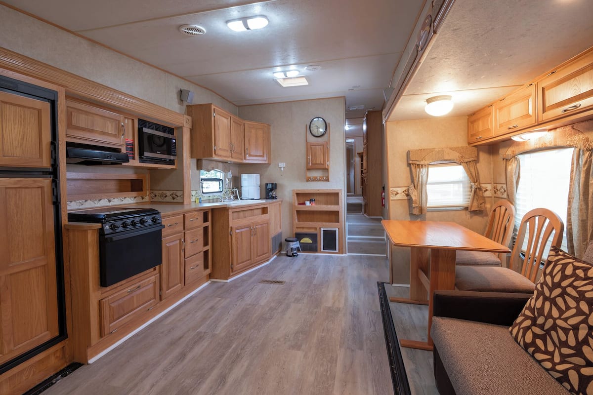 A compact RV kitchen with wooden cabinets, a stove, microwave, and a dining area with a table and chairs.