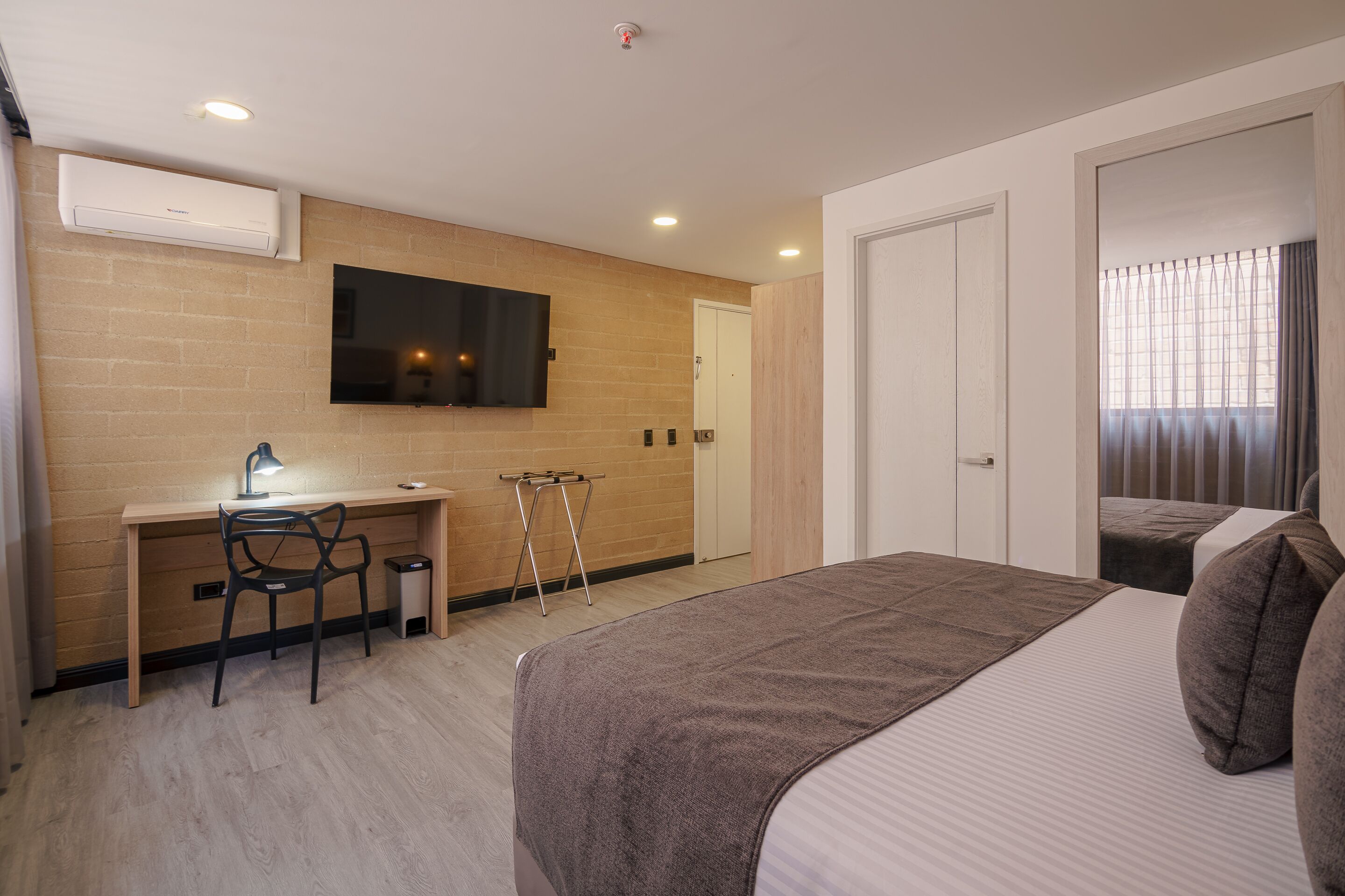 Habitación de hotel con un escritorio de madera, una cama con manta marrón, un televisor montado en la pared y una puerta que da a otra habitación.