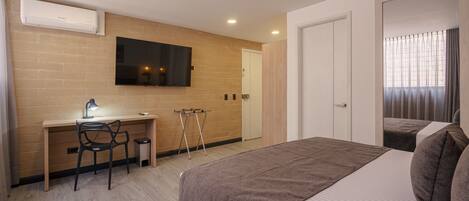 Habitación de hotel con un escritorio de madera, una cama con manta marrón, un televisor montado en la pared y una puerta que da a otra habitación.