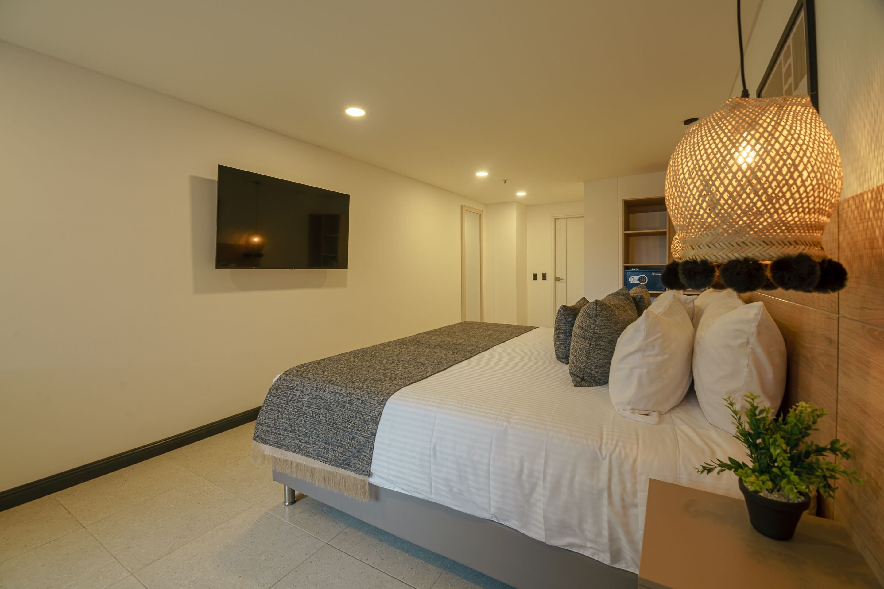 A modern bedroom with a bed, a flat-screen TV, a woven light fixture, and a potted plant.