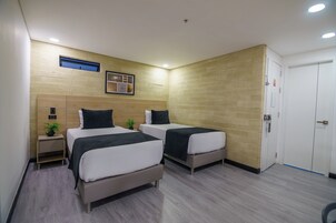 Quarto de hotel com duas camas, uma pequena mesa de cabeceira com uma planta, um respiradouro na parede e uma porta com puxador.