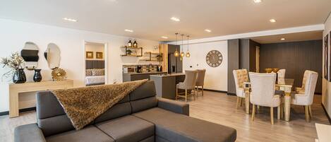 A modern living room with a grey sectional sofa, a dining area with a wooden table and chairs, and a kitchen in the background.