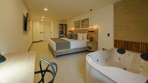 A modern bedroom with a large bed, a sitting area with a chair, a bedside table, a wall-mounted TV, and a large bathtub. - First Class Hotel (Medellín)