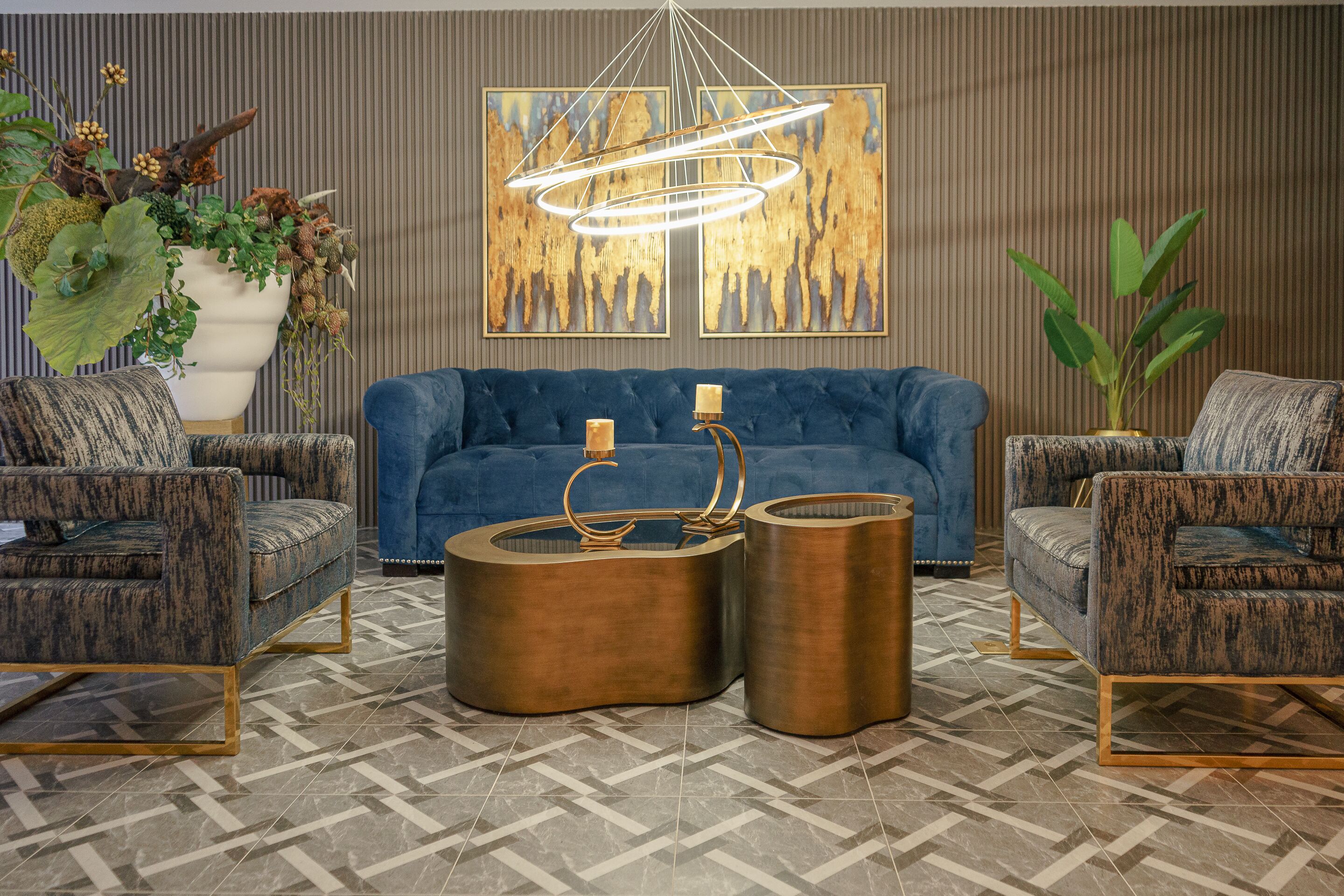 A modern living room with a blue sofa, two patterned armchairs, a large potted plant, a decorative light fixture, and a geometric patterned floor.