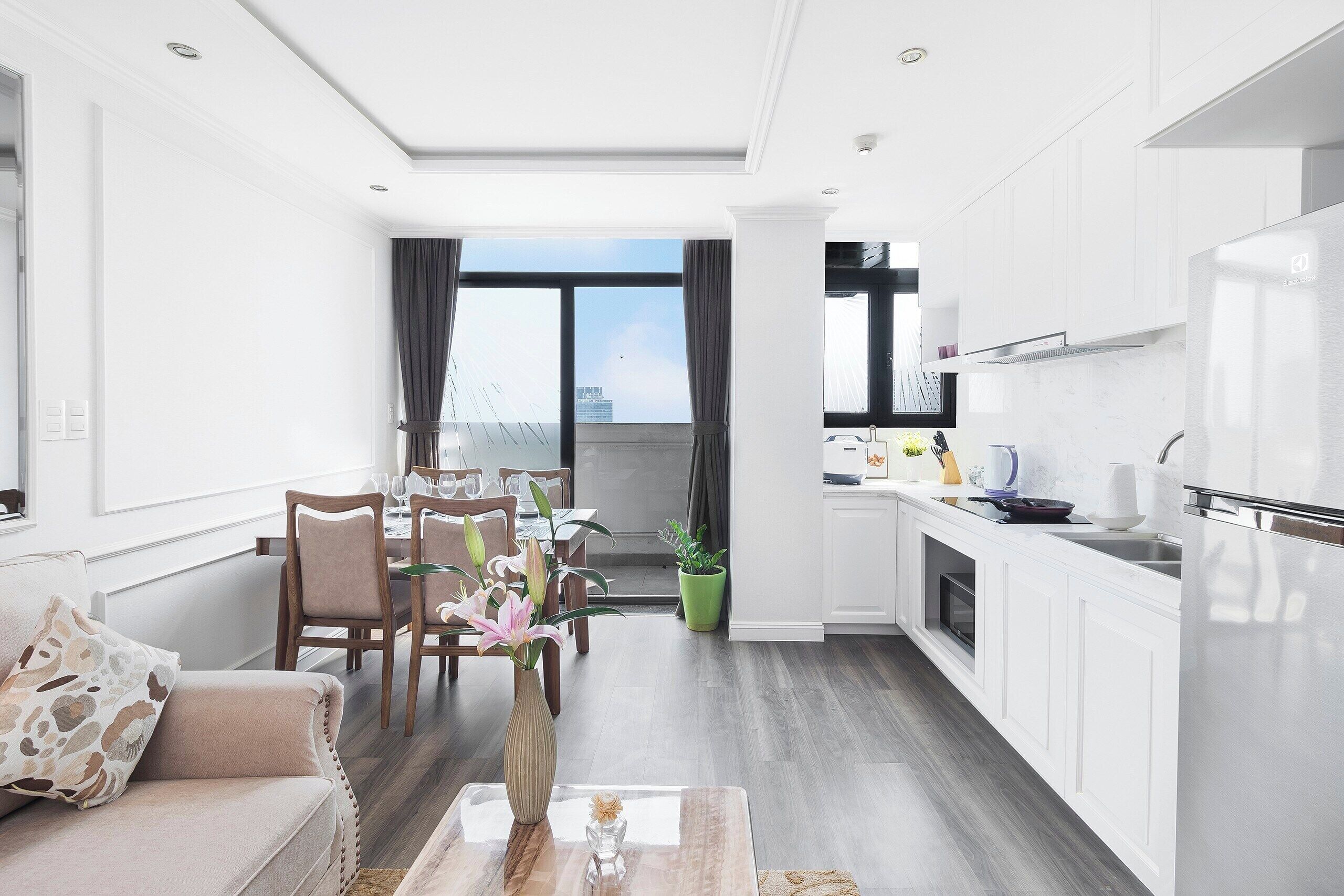 Un salon moderne comprenant un canapé, une table à manger et des chaises, une cuisine avec des meubles blancs et un réfrigérateur, et une vue sur la ville grâce à de grandes fenêtres.