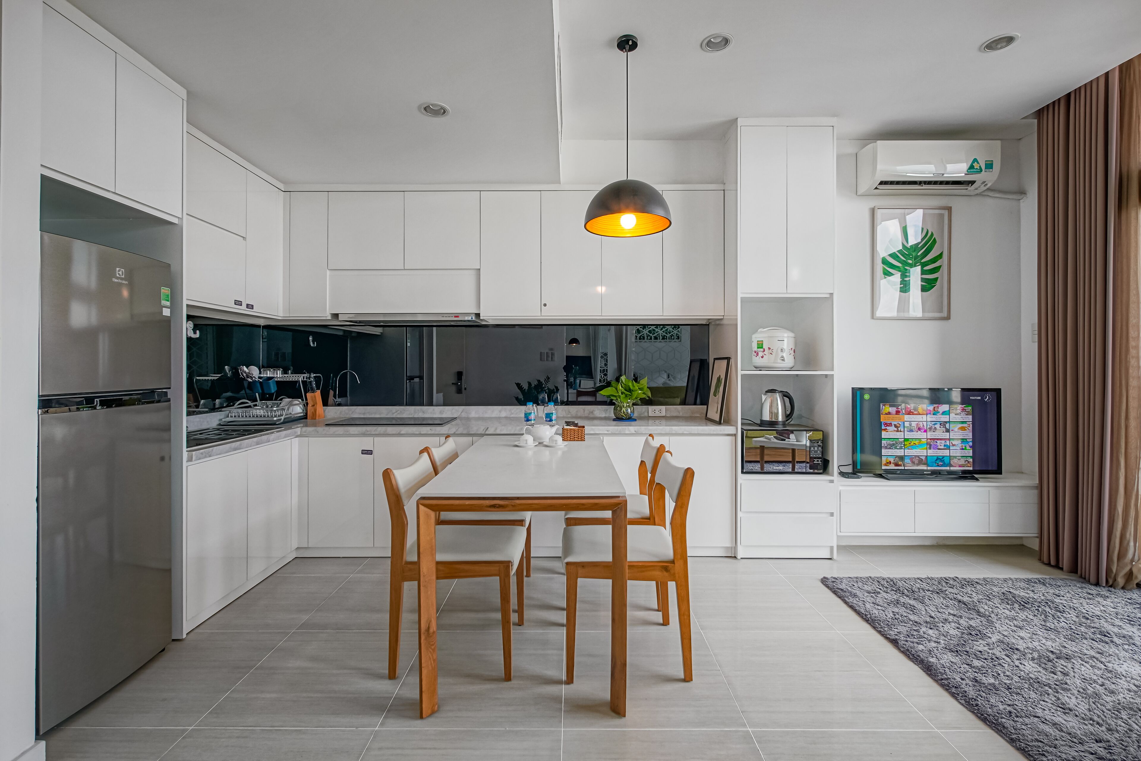 Una cocina moderna con armarios blancos, una isla central con asientos, horno integrado y un televisor en un estante.